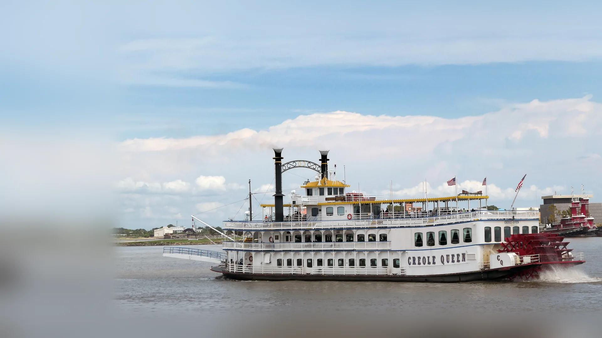 The white paddlewheeler Creole Queen on the Mississippi River, with two black smokestacks, a yellow-trimmed upper deck and a red stern paddle wheel, under a blue sky with a red tugboat behind