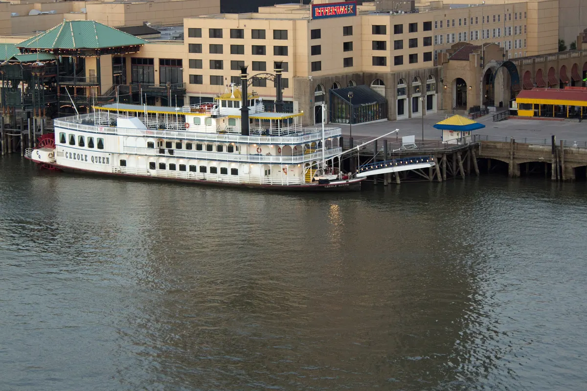 The Creole Queen tied up at the Riverwalk dock in New Orleans, below a row of hotel and shopping buildings, with brown river water in the foreground