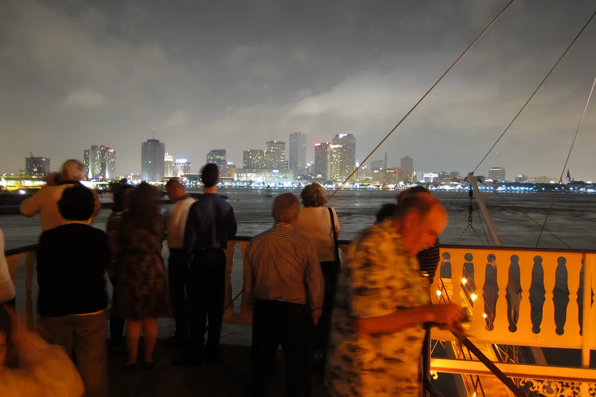 Passengers at the railing of a riverboat deck at night looking across the Mississippi at the lit New Orleans skyline