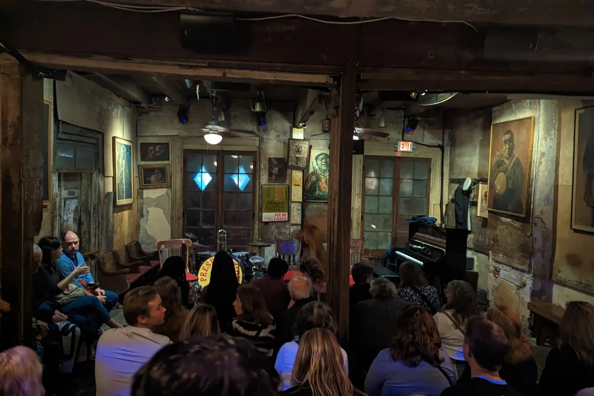 The worn, painted interior of Preservation Hall in the French Quarter with an audience seated facing a small stage and a piano