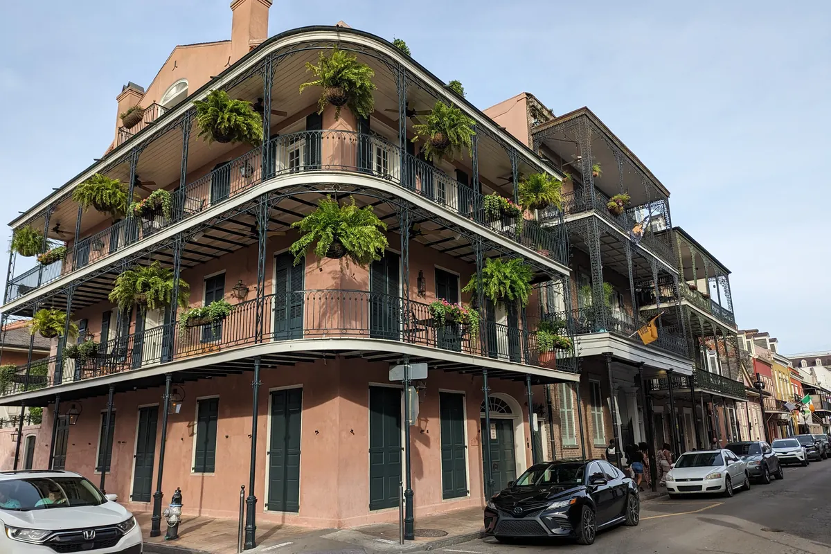 A pink three-storey corner building in the French Quarter with curved cast-iron balconies hung with ferns, and parked cars on the street below