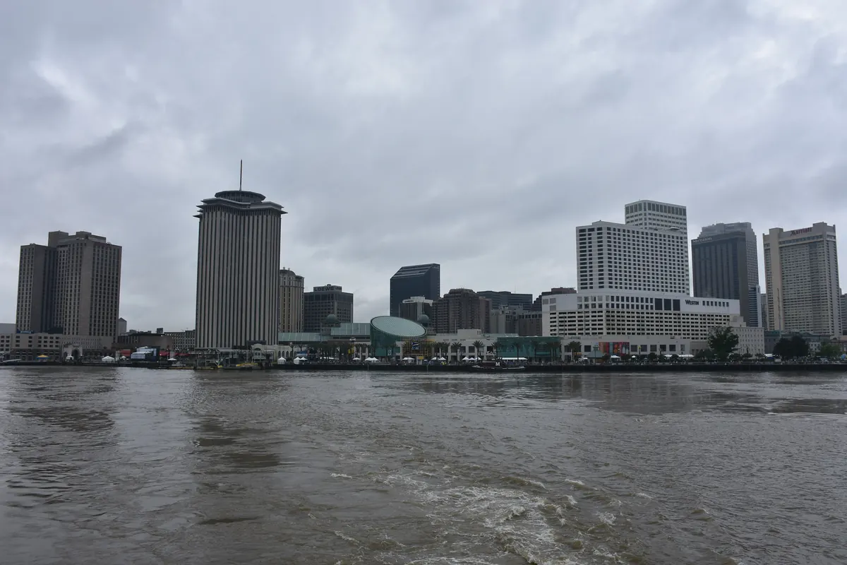 The New Orleans downtown skyline seen across the brown Mississippi River under a grey cloudy sky, with a round-topped tower at the centre