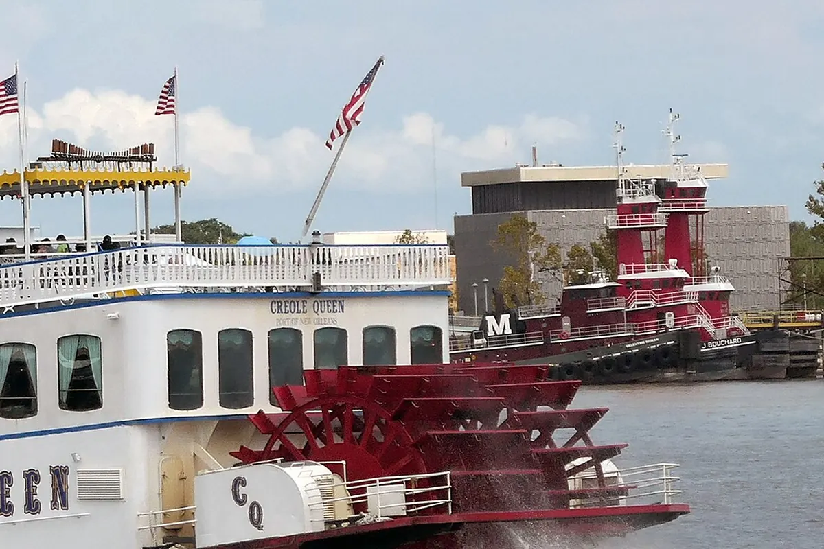 The red stern paddle wheel of the Creole Queen throwing up spray, beside the white lower decks lettered CQ, with a red tugboat behind